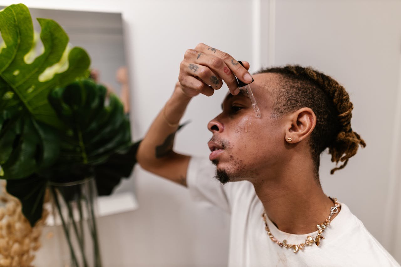 delivery-policy A young man with tattoos and dreadlocks applies skincare serum to his face indoors.